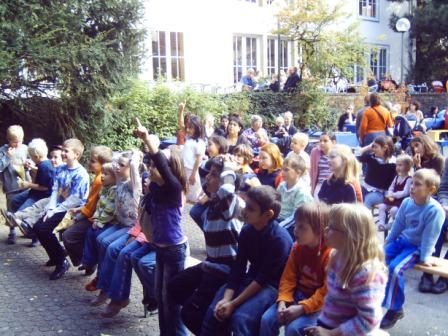 Viele Kinder sitzen im Freien und schauen zu, ein Kind meldet sich.