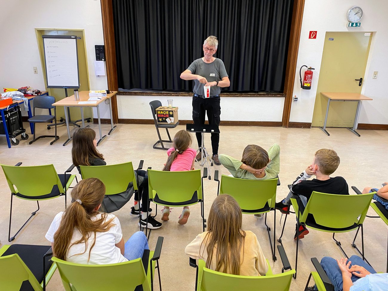 Larifari führt einer Gruppe von Kindern im Halbkreis einen Becher-Trick vor, daneben eine Tafel mit dem Ablauf der Vorstellung.